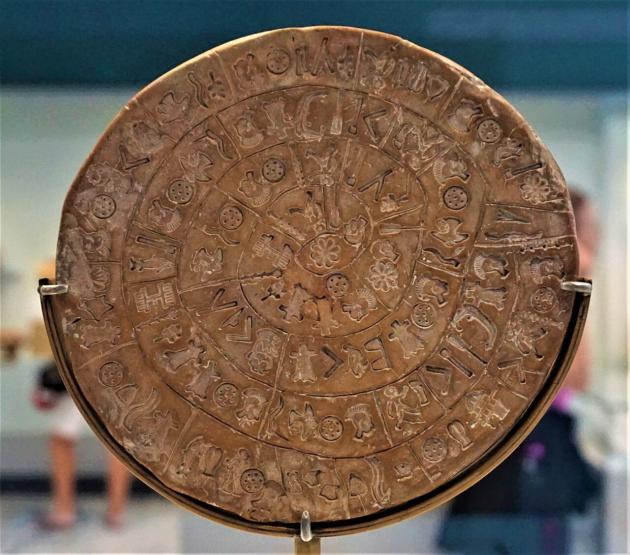 The round clay Phaistos Disc on a metal stand in a museum case, its stamped signs arranged in a spiral of compartments